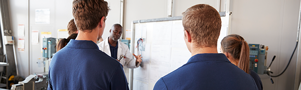 Teacher at a whiteboard talking to apprentices in blue overalls