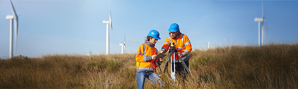 two wind farm engineers using a builder's level to plan out the expansion of the wind farm site. they are wearing orange hi vis jackets and blue hard hats . one is male , one is female. In the background wind turbines can be seen across the landscape.